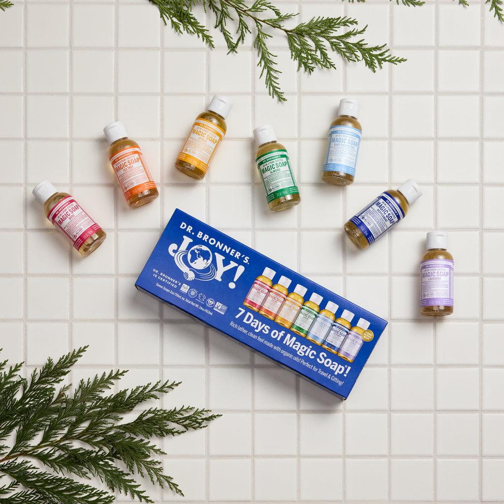 A set of seven small bottles of Dr. Bronners Magic Soap, featuring various colors and scents, surrounds a blue box labeled 7 Days of Magic Soap on a white tiled surface, accented by green pine sprigs [One Size]
