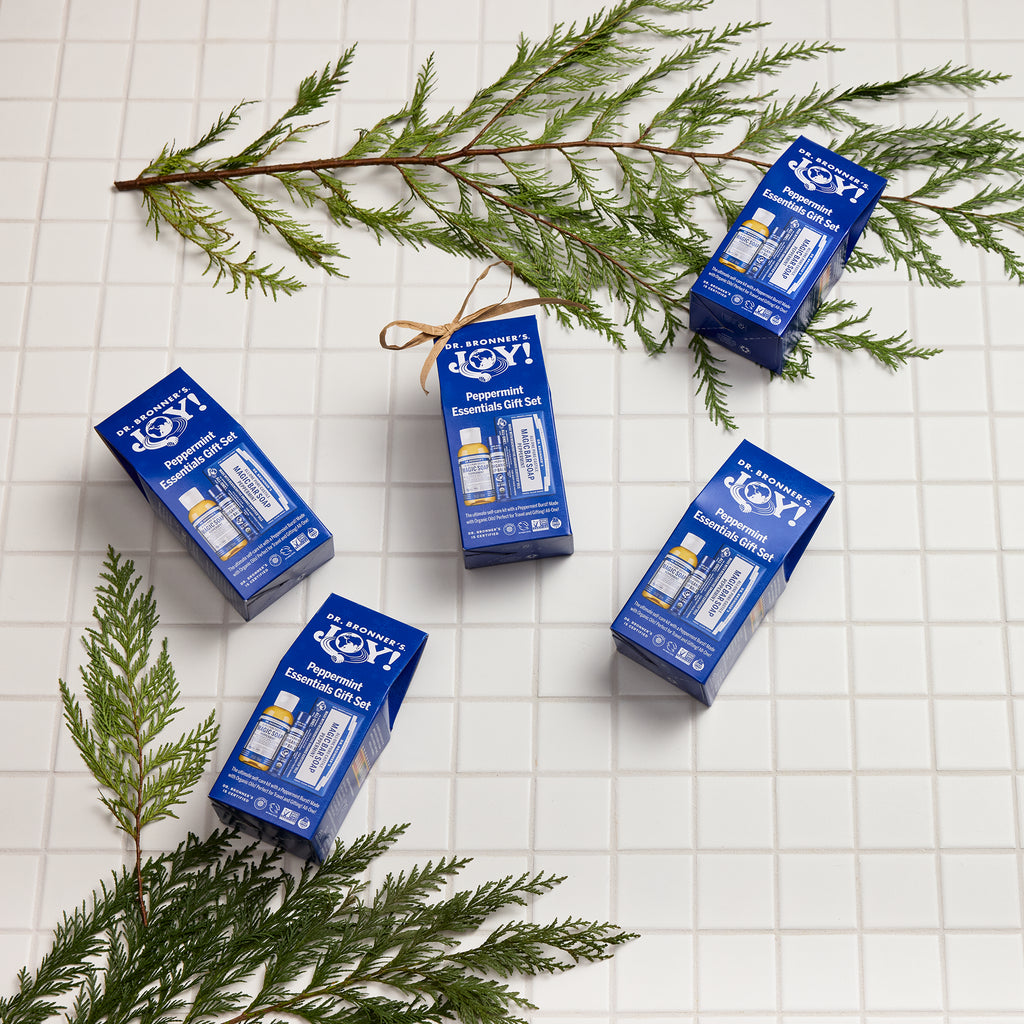 Dr. Bronners peppermint essential gift sets lay on a white tiled surface, surrounded by green pine branches. The packaging highlights Dr. Bronners eco friendly, vegan, organic soap products [One Size]