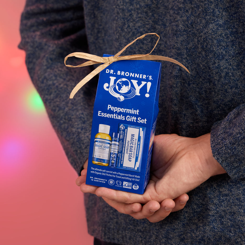 A person holds a blue gift box labeled Dr. Bronners Joy Peppermint Essentials Gift Set, tied with a ribbon, set against a festive, blurred pink background with lights [One Size]