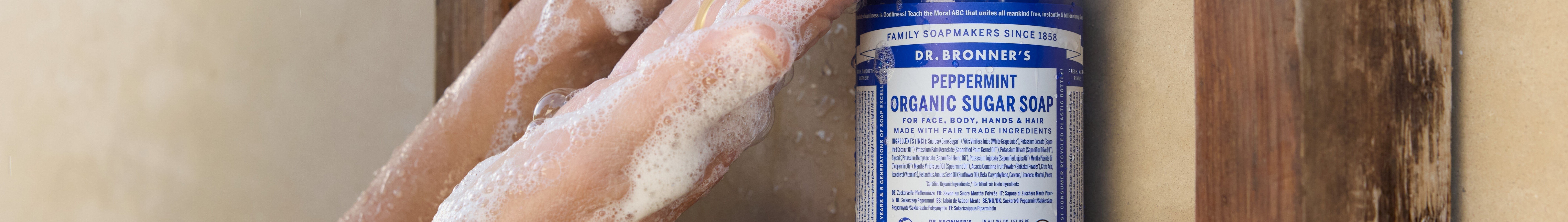 A person holds Dr. Bronners 4 in 1 Peppermint Organic Sugar Soap bottle. Text reads Certified Fair Trade for hands, face, body, hair. Ingredients Organic Sucrose Organic White Grape Juice Organic Coconut Oil Organic Palm Kernel Oil Potassium Hydroxide Organic Olive Oil Organic Shikakai Powder Organic Hemp Oil Organic Jojoba Oil Lime Juice Natural Mentha Arvensis Oil Natural Mentha Piperita Oil Citric Acid Tocopherol. Background bathroom setting. Keywords organic soap, peppermint castile soap