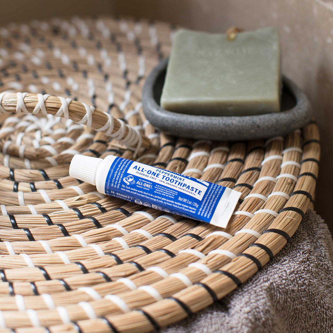 A small tube labeled Dr. Bronners Peppermint Organic Lip Balm lies on a woven mat next to a green bar of soap in a gray dish, creating a natural, eco friendly bathroom scene [1 oz]