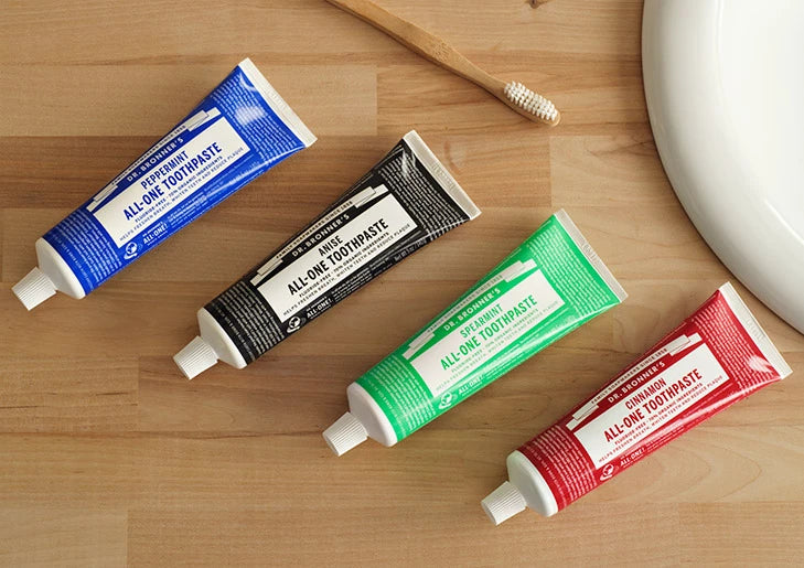 Four toothpaste tubes of various colors lie on a wooden surface next to a toothbrush and a sink. Text reads Dr. Bronners All One Toothpaste [5 oz]