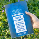A hand holds a book titled HONOR THY Dr. Bronners Journey to a Clean, Green, and Ethical Supply Chain by Gero Leson. The background shows green grass with purple flowers