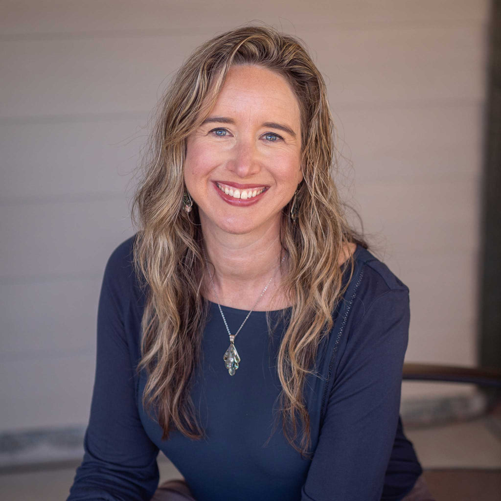 A person with long, wavy hair smiles warmly while sitting indoors. The background features a neutral toned wall, creating a welcoming atmosphere. No text present