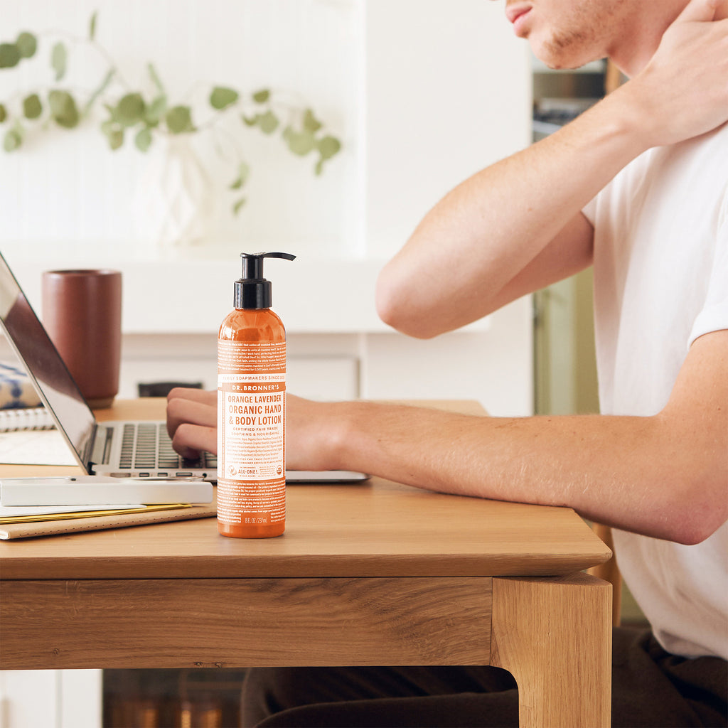 A person sits at a desk using a laptop, massaging their shoulder. A bottle of Dr. Bronners orange labeled liquid soap stands nearby, next to a coffee mug and notebook [8 oz]