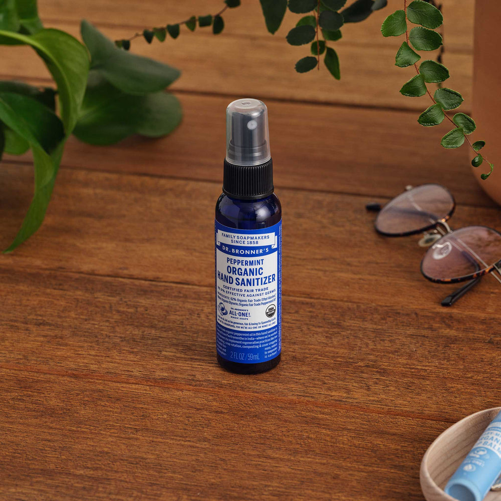 A 2 oz. bottle of Dr. Bronners Peppermint Organic Hand Sanitizer stands on a wooden surface near green leaves and sunglasses, emphasizing natural, eco friendly hygiene. Dr. Bronners Peppermint Organic Hand Sanitizer