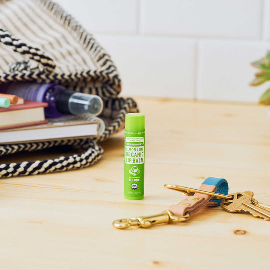 A lemon lime organic lip balm stands upright on a wooden surface beside a set of keys with colorful keychains. In the background, a patterned blanket and books create a cozy atmosphere [.15 oz]