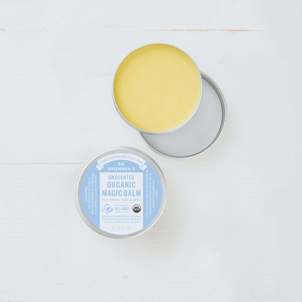 A round tin container with its lid off reveals a yellow balm inside. The label reads, Dr. Bronners Magic Balm, Baby Unscented. Its set on a light surface  [2 oz]