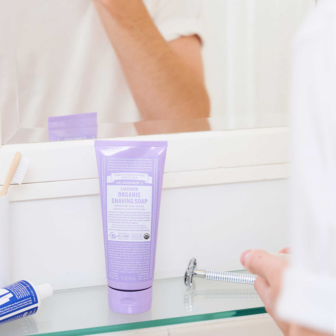 A lavender tube labeled Dr. Bronners sits on a bathroom sink beside a toothbrush and a razor, with a person reflected in the mirror above, suggesting a morning routine [7 oz]