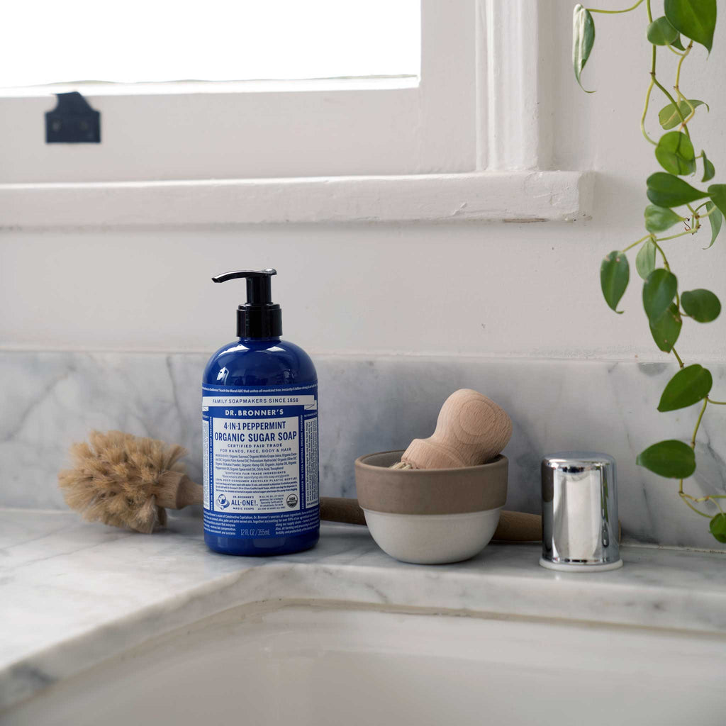 A blue bottle of Dr. Bronners liquid castile soap sits on a bathroom counter near a sink. A natural brush and a small plant add an eco friendly touch to the clean and serene setting [12 oz]
