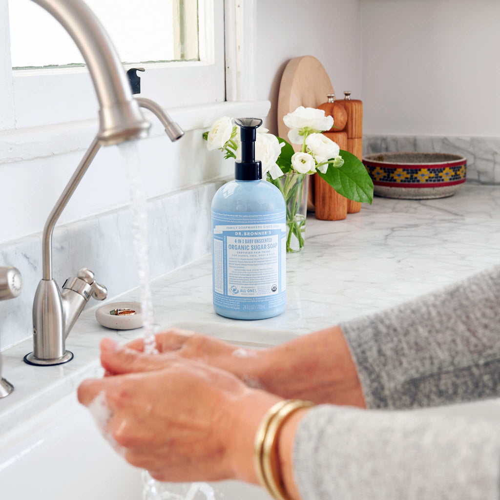 Hands lather with Dr. Bronners Organic Sugar Soap under a kitchen faucet, set against a bright countertop with decorative elements like flowers and dishes nearby [24 oz]