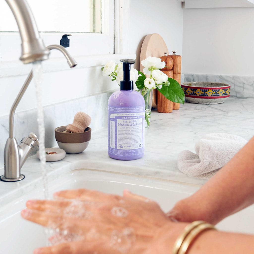 Hands are being washed under a running tap with Dr. Bronners 4 in 1 Lavender Organic Sugar Soap on a marble counter. Nearby, a plant, bowl, and towel enhance the bathroom decor [24 oz]