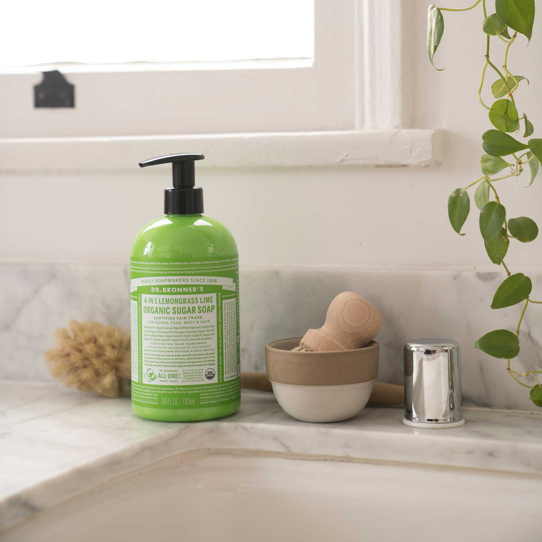 Dr. Bronners Lemongrass Lime Organic Sugar Soap bottle sits on a marble bathroom counter near a small bowl, wooden brush, and leafy plant, providing natural and eco friendly skincare options [24 oz]