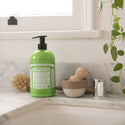 Dr. Bronners Lemongrass Lime Organic Sugar Soap bottle sits on a marble bathroom counter near a small bowl, wooden brush, and leafy plant, providing natural and eco friendly skincare options [24 oz]