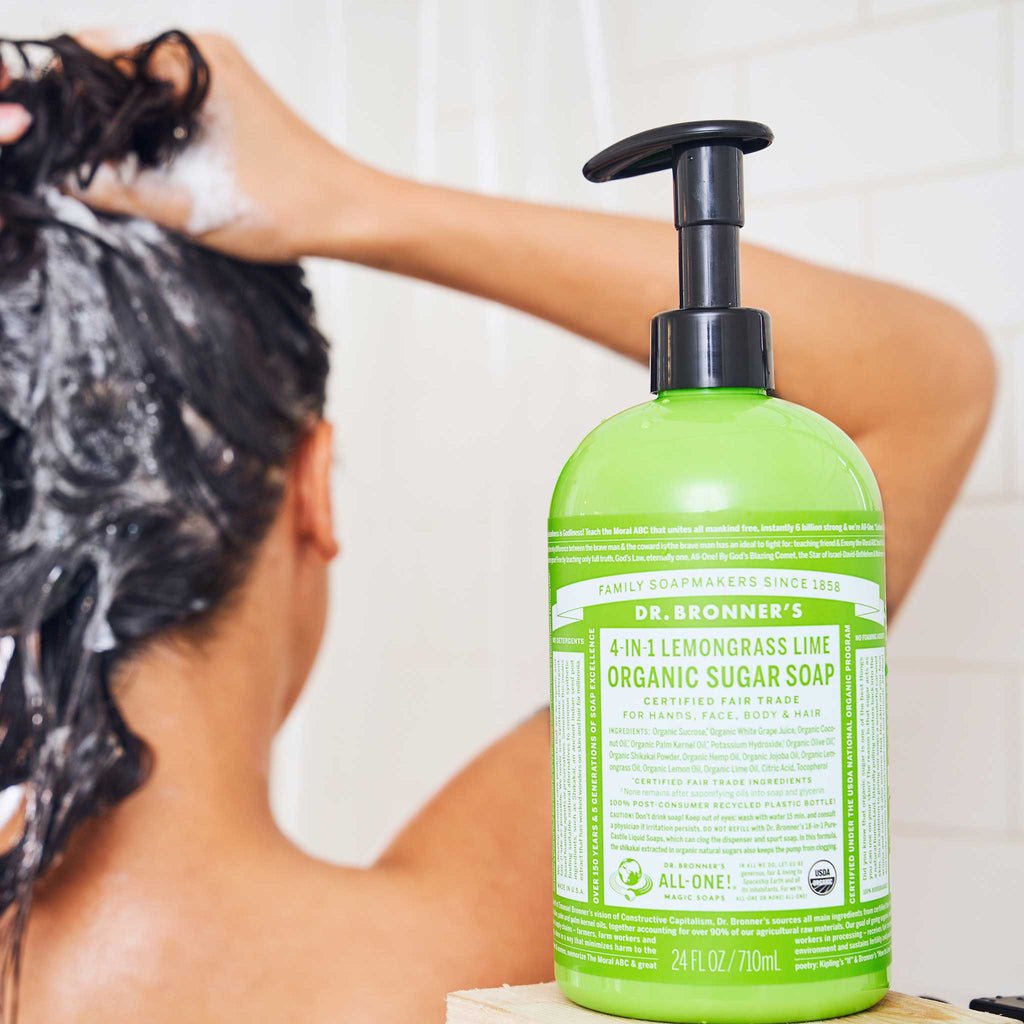A person shampoos their hair in a shower, with a bottle of Dr. Bronners 4 in 1 Lemongrass Lime Organic Sugar Soap in the foreground. The green bottle emphasizes its versatility for hair, face, body, and hands [24 oz]