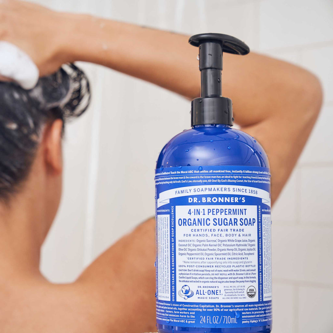 A person washes their hair in a shower using a blue bottle of Dr. Bronners 4 in 1 Peppermint Organic Sugar Soap, which is certified fair trade for hands, face, body, and hair [24 oz]