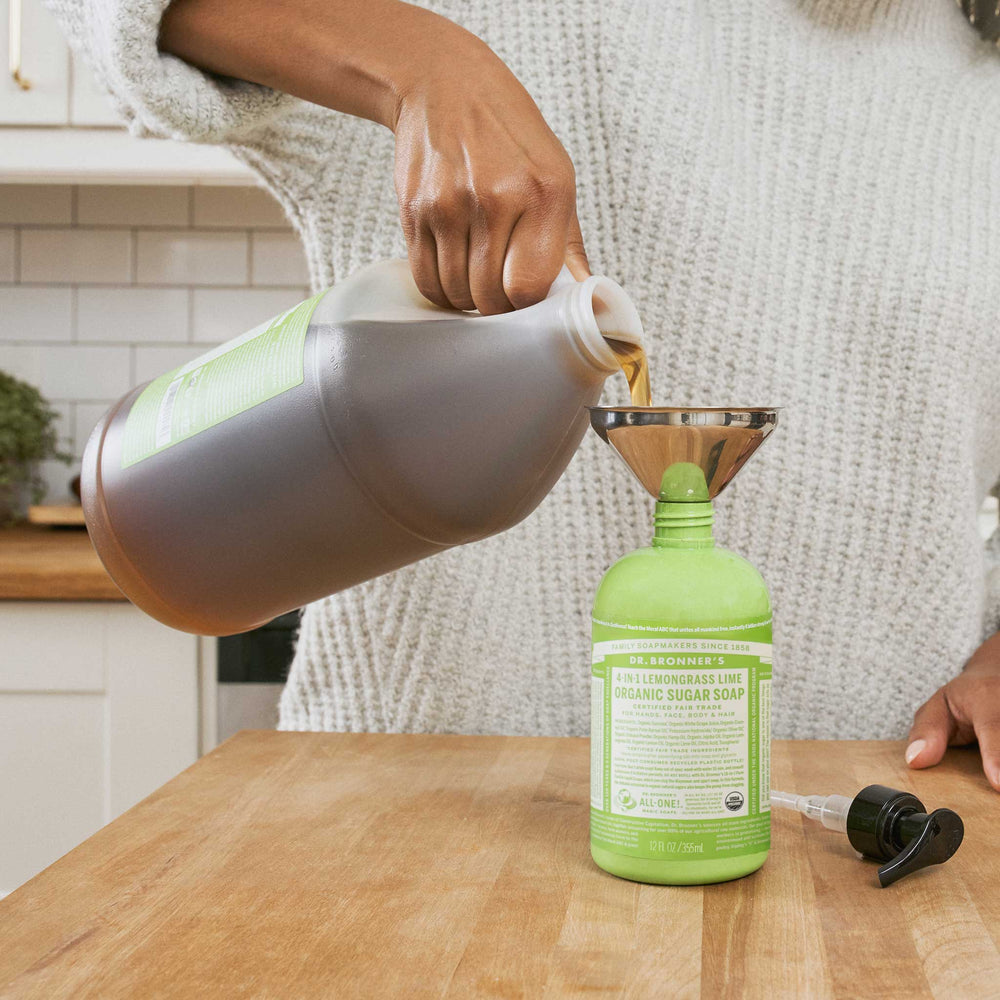 A person pours Dr. Bronners Lemongrass Lime Organic Sugar Soap from a large container into a smaller bottle using a funnel, in a kitchen setting [12 oz]