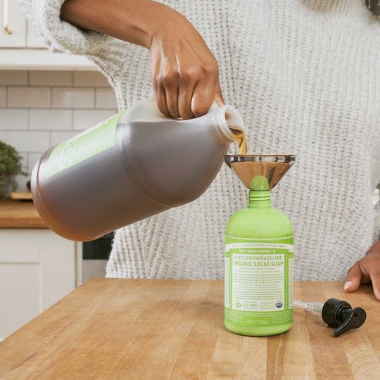 A person pours Dr. Bronners Lemongrass Lime Organic Sugar Soap from a large container into a smaller bottle using a funnel, in a kitchen setting [12 oz]