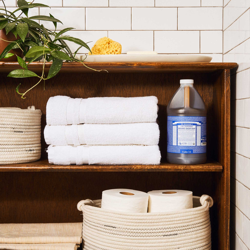 A bottle of Dr. Bronners liquid castile soap sits on a wooden bathroom shelf beside neatly folded white towels. Nearby, wicker baskets hold toilet paper and a potted plant enhances the natural, organized setting [1/2 Gallon]