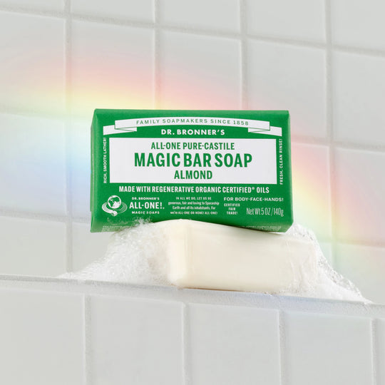 Dr. Bronners Magic Bar Soap in almond scent sits on a soap dish, generating bubbles against a tiled wall with a visible rainbow reflection. FAMILY SOAPMAKERS SINCE 1858, ALL ONE PURE CASTILE MAGIC BAR SOAP ALMOND, MADE WITH REGENERATIVE ORGANIC CERTIFIED OILS, FOR BODY FACE HANDS CERTIFIED FAIR TRADE [5 oz]