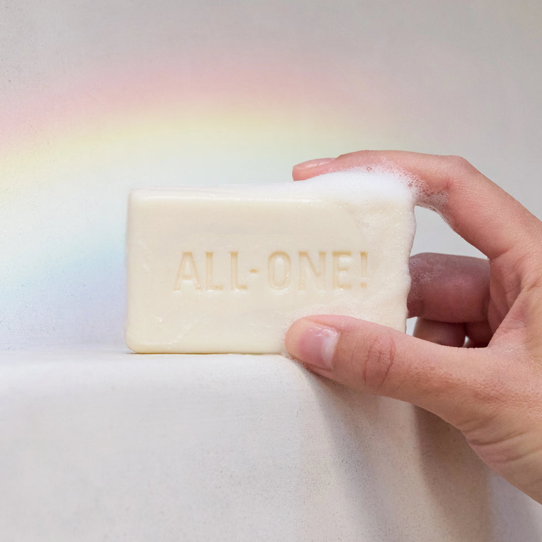 A hand holds a bubbly bar of castile soap embossed with ALL ONE against a light background with a subtle rainbow. Ideal for promoting Dr. Bronners natural, eco friendly products [5 oz]