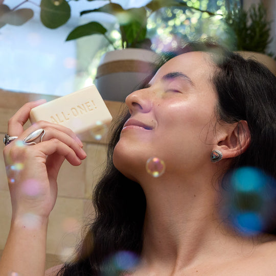 A woman with dark hair holds a DR BRONNERS ALL ONE bar castile soap near her face with a content expression, surrounded by bubbles in a light filled bathroom adorned with plants [5 oz]