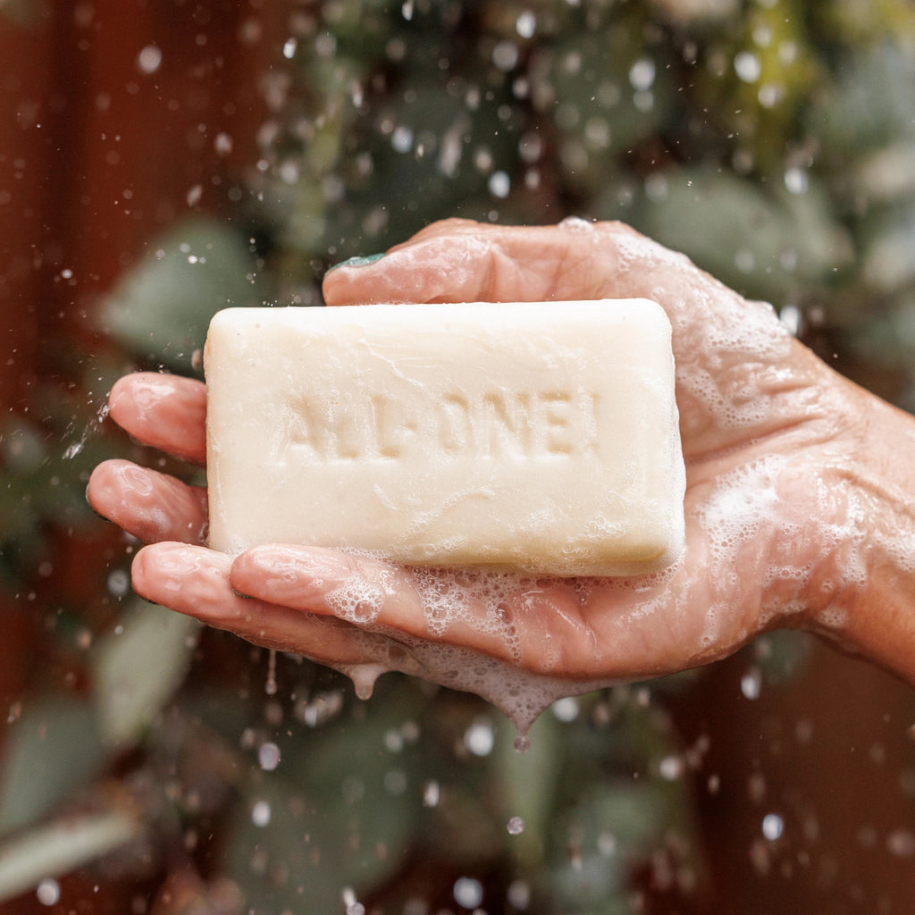 A hand holds a sudsy bar of soap with ALL ONE text embossed, against a blurred leafy background. This captures the essence of natural, vegan, eco friendly soap choices [5 oz]