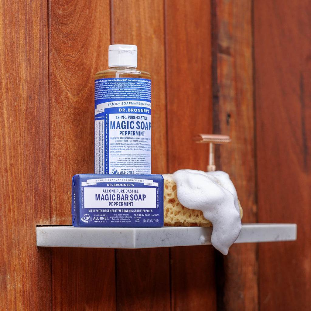 A bottle of Dr. Bronners Pure Castile Liquid Soap and a bar of soap sit on a shower shelf, surrounded by wood panel walls. Foam spills over an attached loofah, emphasizing cleanliness [5 oz]