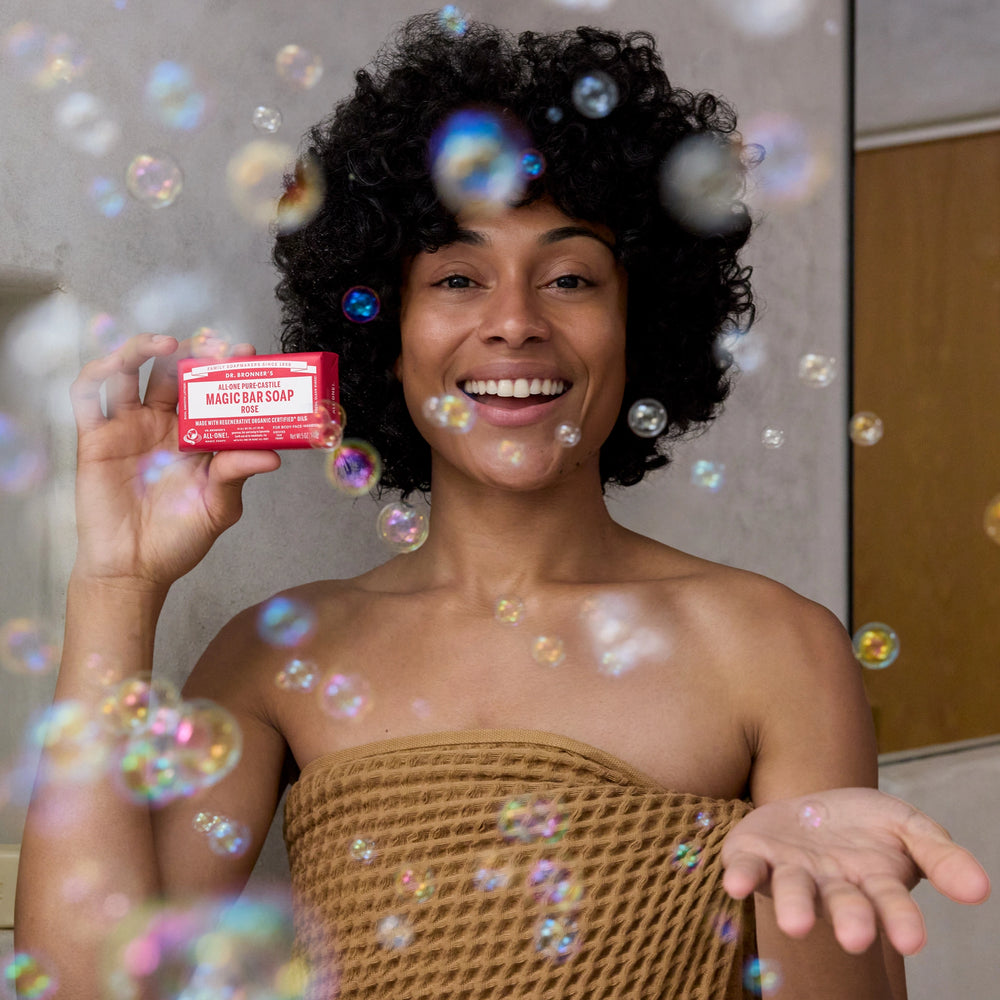 A person holds a red Dr. Bronners bar soap in one hand and smiles, surrounded by floating bubbles, in a bathroom setting, wearing a checkered towel [5 oz]