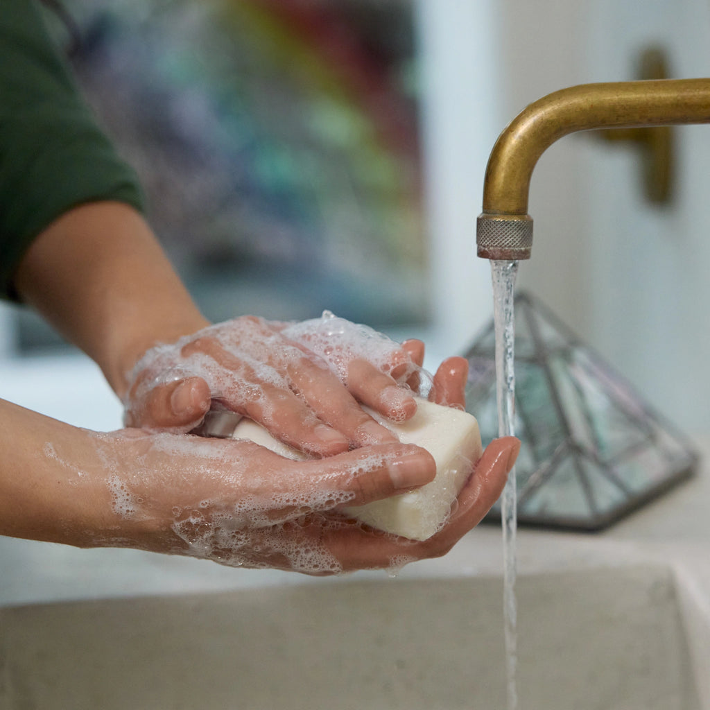 Hands lather a bar of soap under running water from a brass faucet, positioned above a kitchen sink. The background features a blurred, colorful wall, creating a cozy ambiance [5 oz]