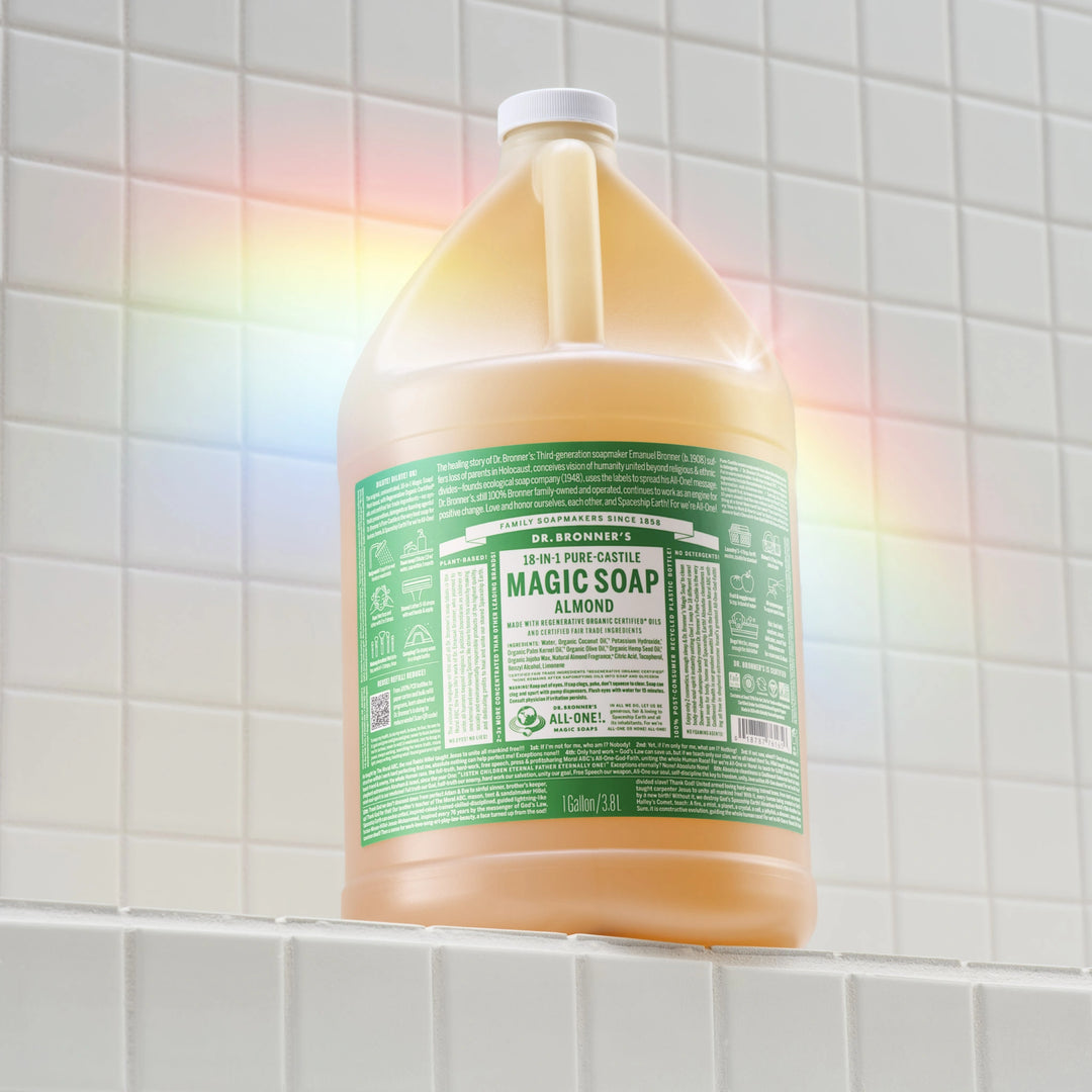 A gallon jug of Dr. Bronners almond castile soap stands on a tiled surface, illuminated by a rainbow light. Text on the label includes DR. BRONNERS 18 IN 1 PURE CASTILE MAGIC SOAP ALMOND [1 Gallon]