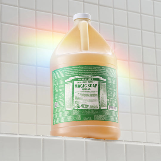 A gallon jug of Dr. Bronners almond castile soap stands on a tiled surface, illuminated by a rainbow light. Text on the label includes DR. BRONNERS 18 IN 1 PURE CASTILE MAGIC SOAP ALMOND [1 Gallon]