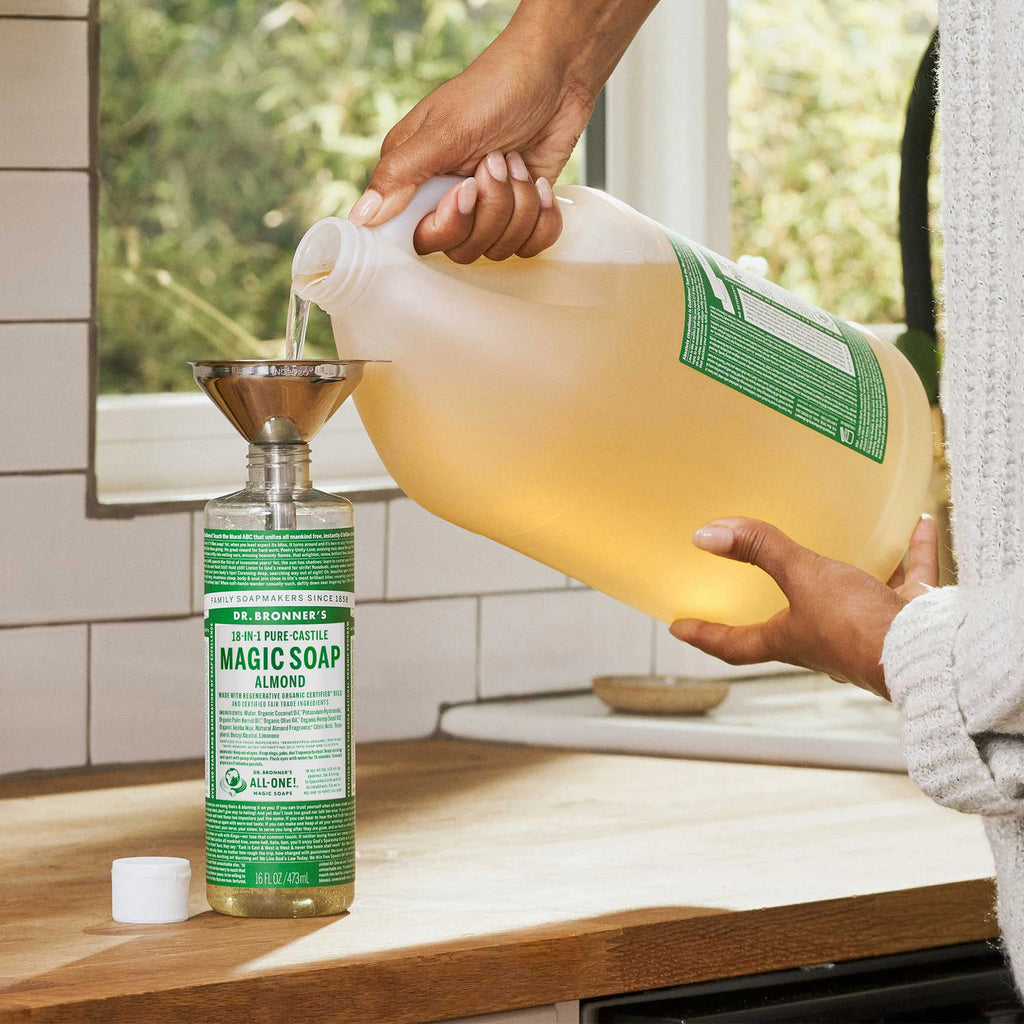 A hand pours Dr. Bronners liquid castile soap from a large jug into a smaller bottle using a funnel on a wooden kitchen countertop, near a window with greenery outside [16 oz, 1 Gallon]