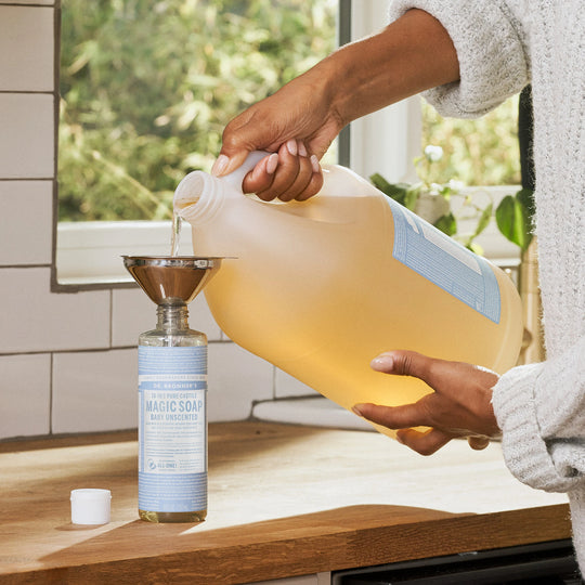 A person pours Dr. Bronners Baby Unscented Pure Castile Liquid Soap from a large bottle into a smaller bottle using a funnel on a kitchen counter, creating an eco friendly refill setting [16 oz, 1 Gallon]