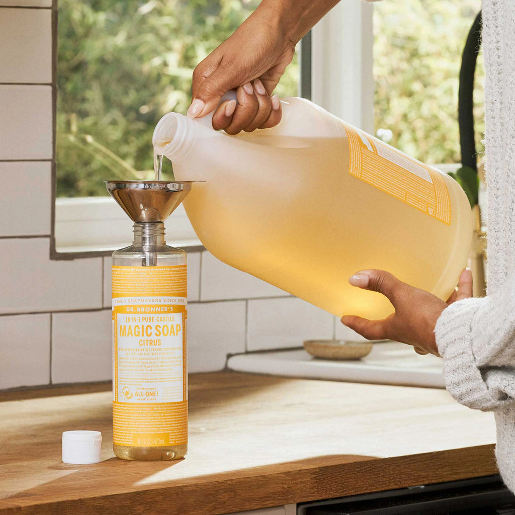 A large jug is pouring liquid soap into a smaller bottle via a funnel on a kitchen counter. The window behind suggests a well lit, natural setting [16 oz, 1 Gallon]