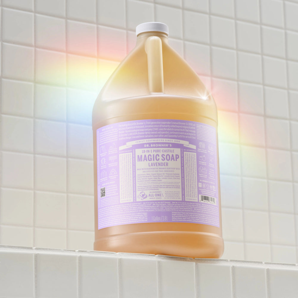 A large bottle of Dr. Bronners Lavender Pure Castile Soap sits on a tiled bathroom shelf, reflecting a rainbow light. The lavender label details its vegan, organic, and versatile cleaning uses [1 Gallon]