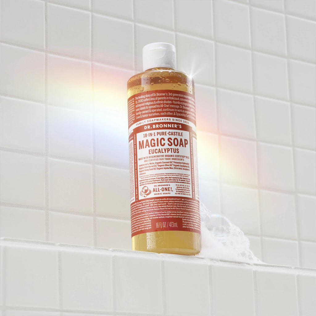 A bottle of Dr. Bronners 18 in 1 Pure Castile Magic Soap in eucalyptus scent rests on a white tiled bathroom shelf, surrounded by colorful light reflections and suds. Text reads DR. BRONNERS 18 IN 1 PURE CASTILE MAGIC SOAP EUCALYPTUS. Made with regenerative organic certified oils and certified fair trade ingredients for the betterment of people and planet [16 oz]