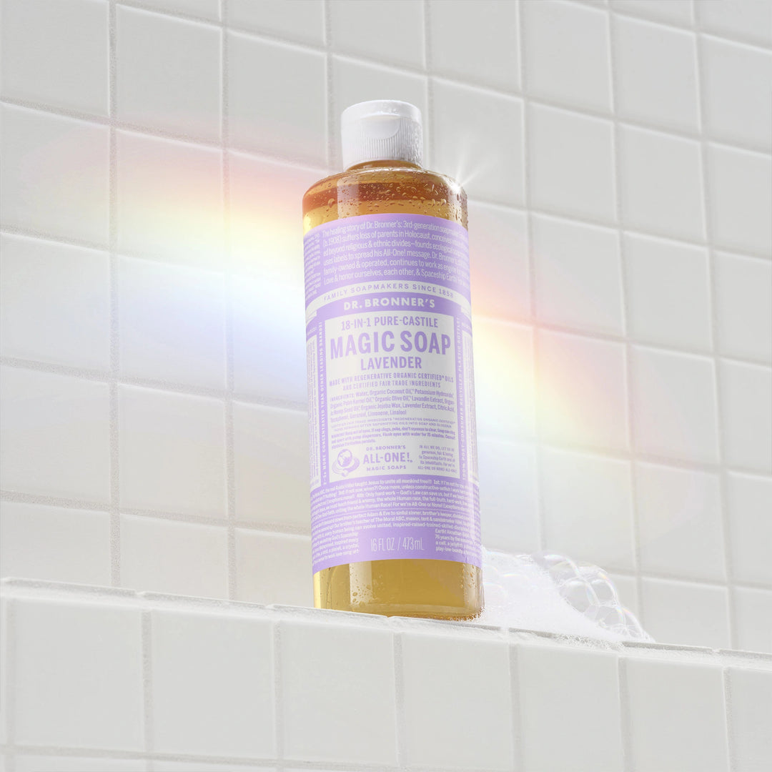 A bottle of Dr. Bronners lavender liquid castile soap sits on a tiled shower shelf, illuminated by a soft rainbow light. The label features text highlighting its organic, eco friendly ingredients [16 oz]