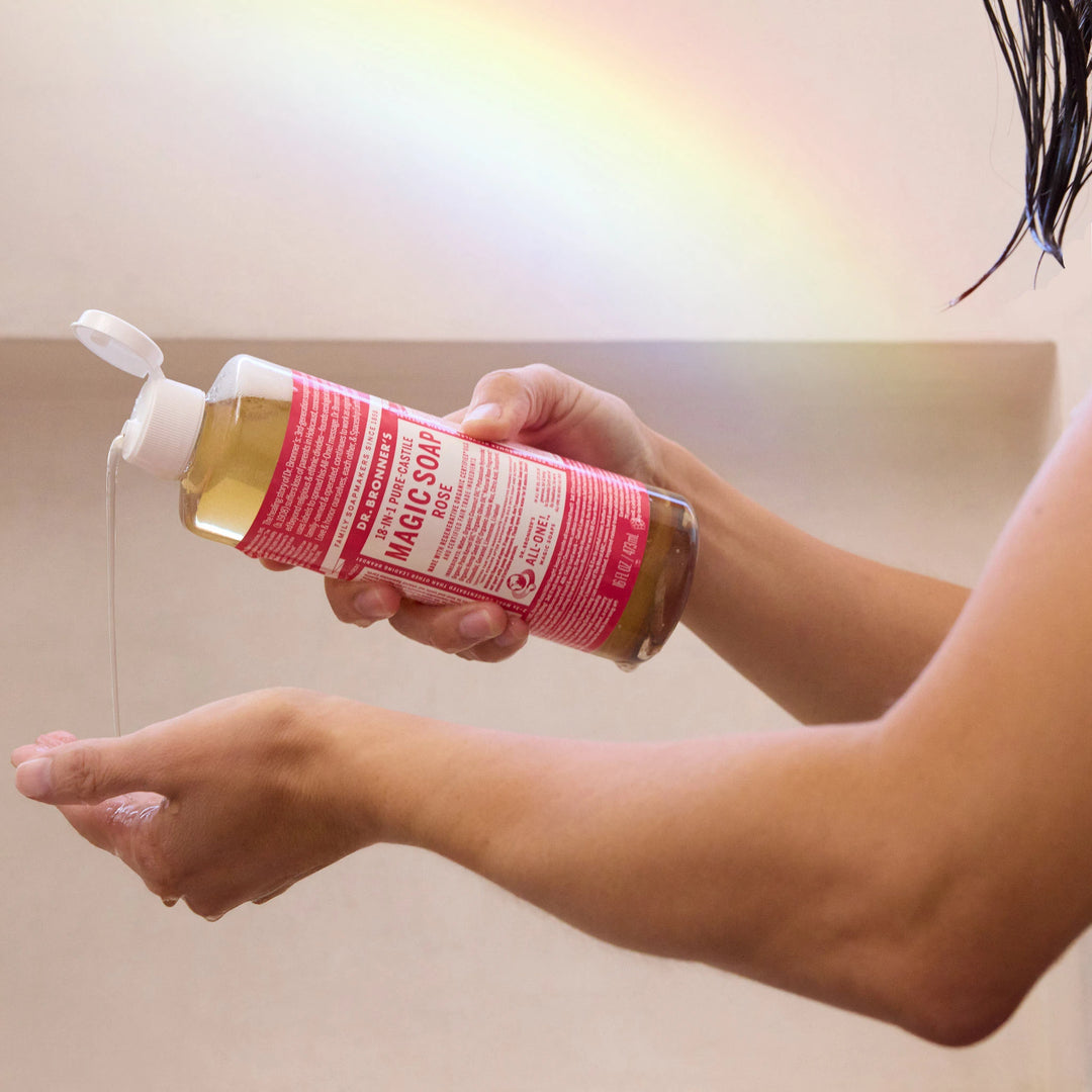 A person dispenses liquid from a pink bottle of Dr. Bronners pure castile soap into their palm, under a soft rainbow light in a bathroom setting [16 oz]