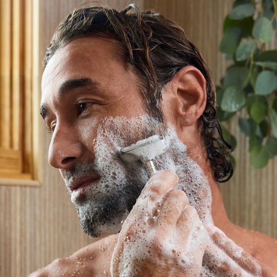 A man with wet hair and a foamy beard shaving his cheek with a razor in a bathroom setting. [16 oz]