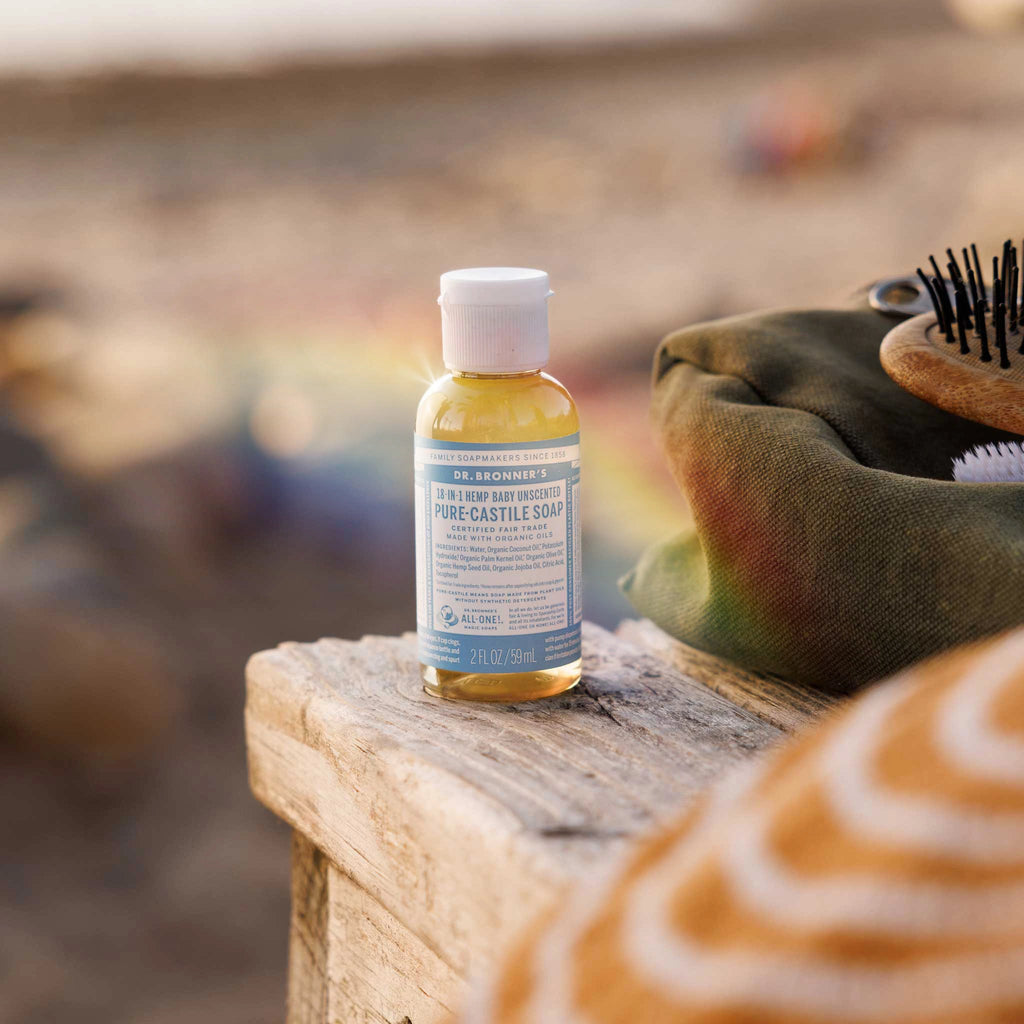 A bottle of Dr. Bronners 18 in 1 Hemp Baby Unscented Pure Castile Soap rests on a wooden surface, surrounded by a bag and brush, with a blurred outdoor background [2 oz]