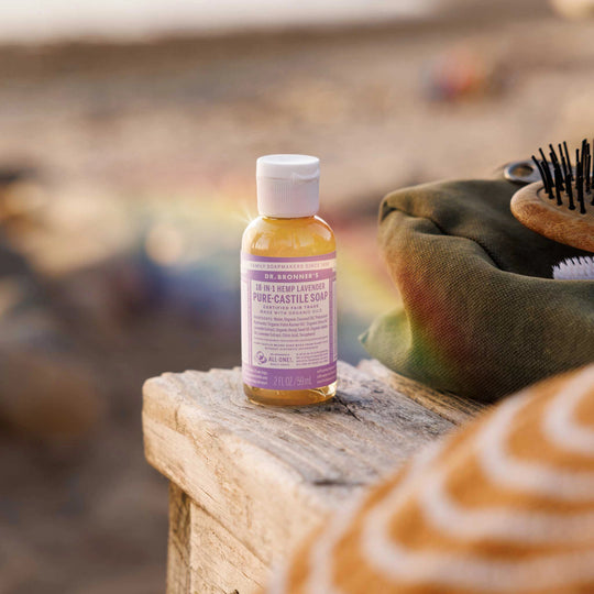 A small bottle of Dr. Bronners 18 in 1 Hemp Lavender Pure Castile Soap rests on a wooden surface at a beach. Nearby, a colorful bag and a brush add context [2 oz]