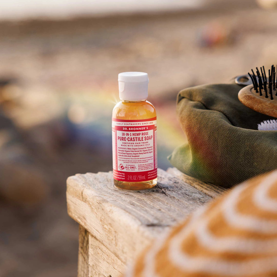 A small bottle of Dr. Bronners peppermint castile soap sits on a wooden surface outdoors, surrounded by a soft focus beach setting. The label is prominently displayed [2 oz]