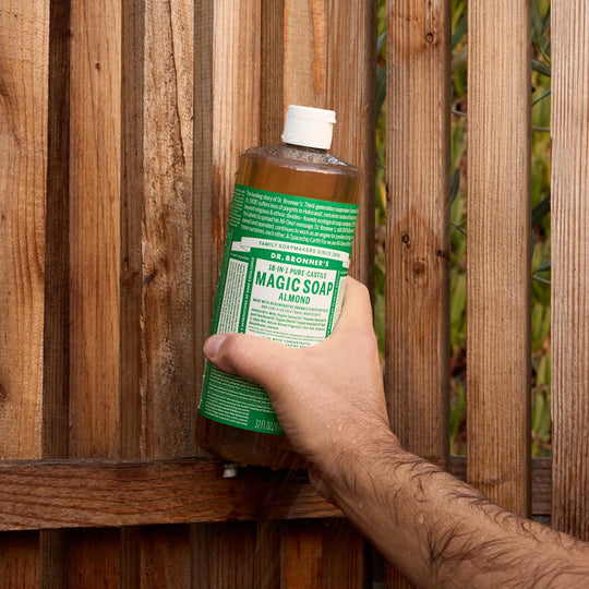 A hand holds a bottle of Dr. Bronners liquid castile soap against a wooden fence. The green label suggests it might be the eucalyptus castile soap. The setting is outdoors [32 oz]