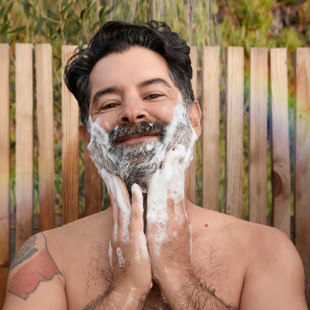 A person lathers soap on their face, smiling contently, in front of a wooden fence with greenery behind. Perfect for showcasing the benefits of Dr. Bronners organic, vegan, and sustainable castile soap [2 oz, 3.4 oz, 4 oz, 8 oz, 16 oz, 32 oz, 1 Gallon]