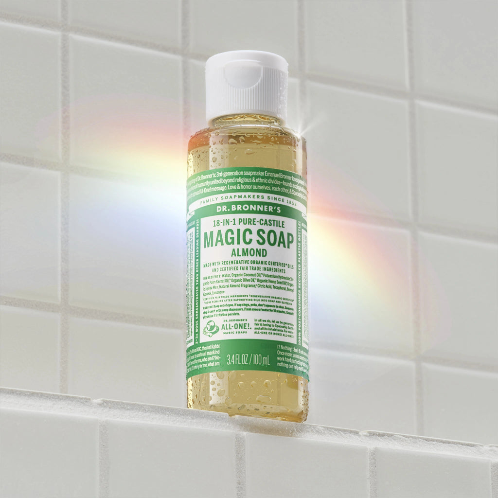 A bottle of Dr. Bronners Magic Soap with a green label, floating against white tiled background, casts a rainbow reflection. Text on bottle Magic Soap, Pure Castile Soap, 18 in 1 Hemp [3.4 oz]