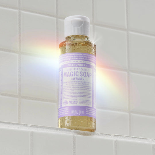 A small bottle of Dr. Bronners Pure Castile Magic Soap in lavender scent stands on a tiled surface, illuminated by a rainbow light. The label reads DR. BRONNERS 18 IN 1 PURE CASTILE MAGIC SOAP LAVENDER [3.4 oz]