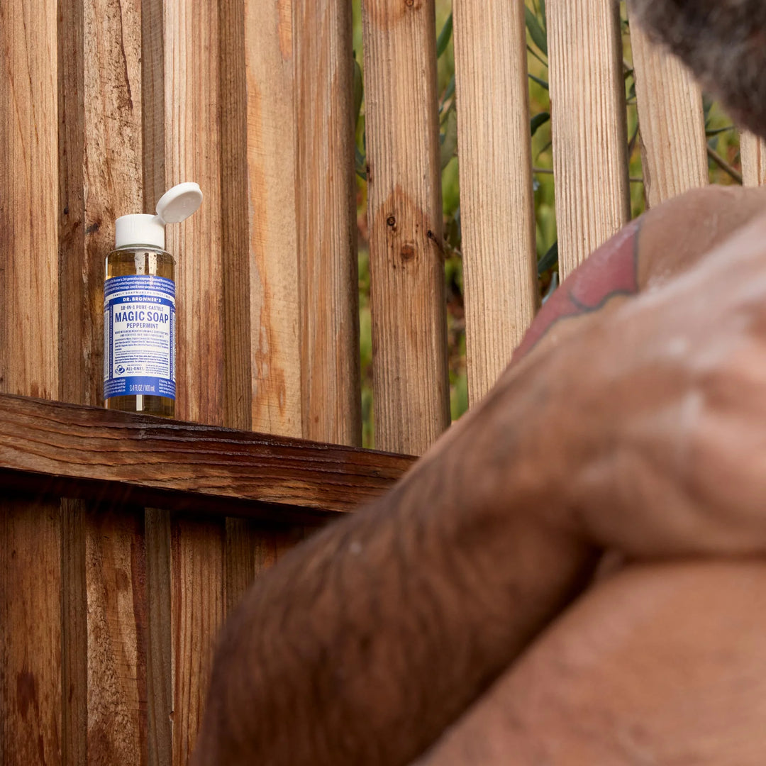 A bottle of Dr. Bronners Magic Soap in peppermint flavor sits on a wooden shelf in an outdoor shower, with a person washing nearby, surrounded by a wooden fence [3.4 oz]
