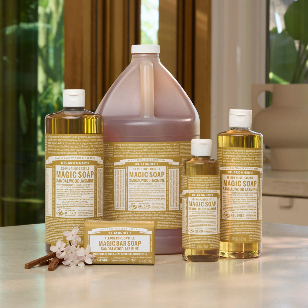 A set of Dr. Bronner's Magic Soap sandalwood jasmine liquid and bar soaps arranged on a light countertop with a blurred kitchen background. [3.4 oz, 8 oz, 16 oz, 32 oz, 1 Gal]