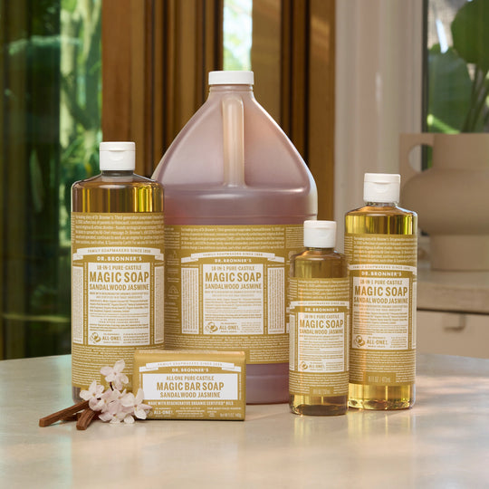 A set of Dr. Bronner's Magic Soap sandalwood jasmine liquid and bar soaps arranged on a light countertop with a blurred kitchen background. [3.4 oz, 8 oz, 16 oz, 32 oz, 1 Gal]